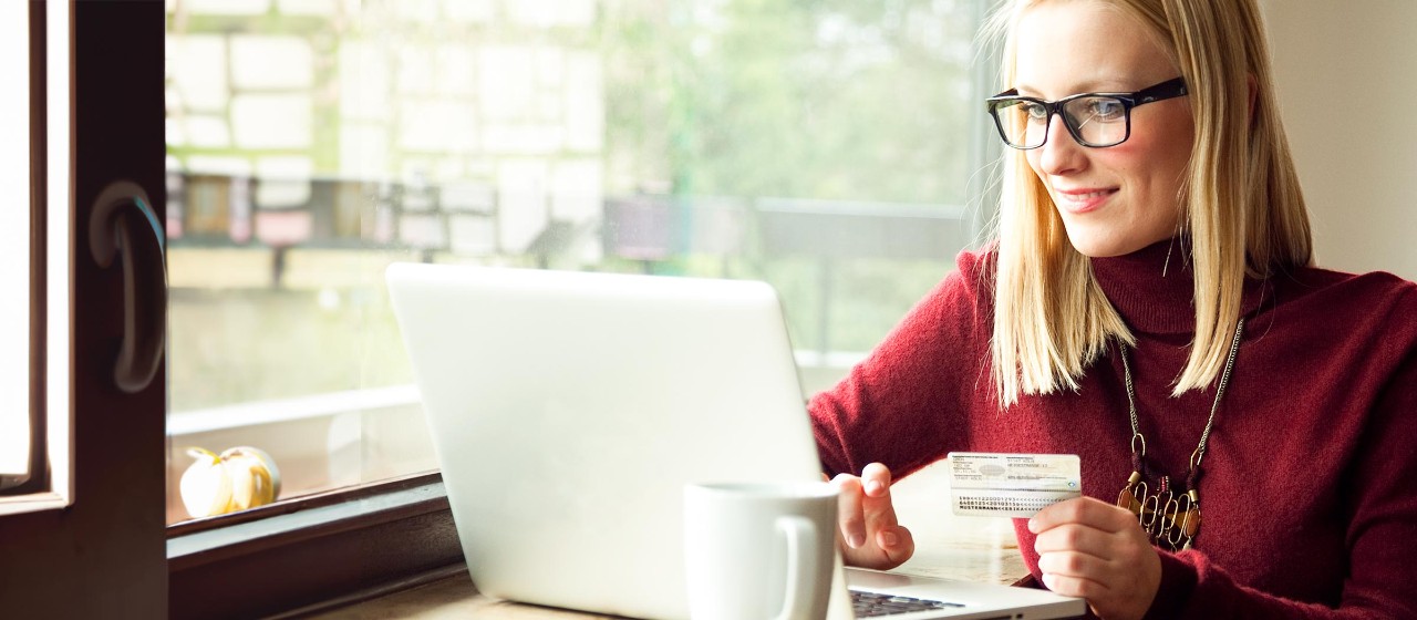 Frau mit schulterlangen blonden Haaren, dunkelrotem Pullover und schwarzer Brille mit ihrem Ausweis vor Ihrem Laptop
